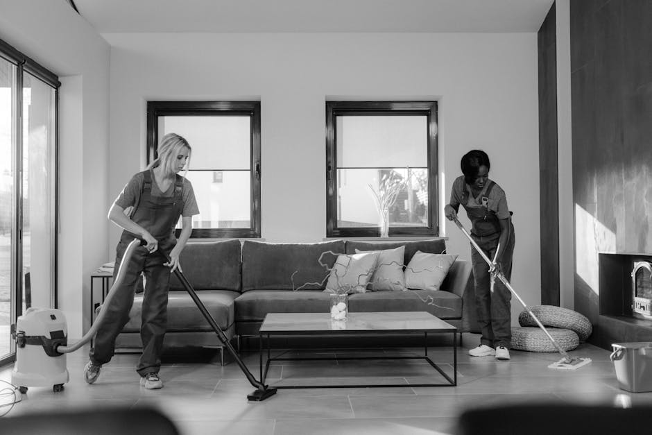 A person wearing a blue disposable glove is wiping a black remote control with a blue cleaning cloth, placed on a smooth, light-colored surface in a domestic setting. The remote control features vario