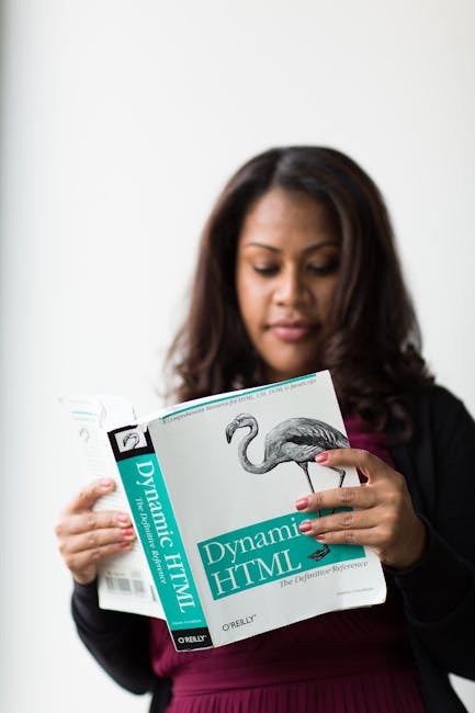 A woman with dark brown, shoulder-length hair, dressed in a black blazer over a maroon top, is holding an open book titled 'Dynamic HTML' by O'Reilly. She is examining the pages carefully, with her li