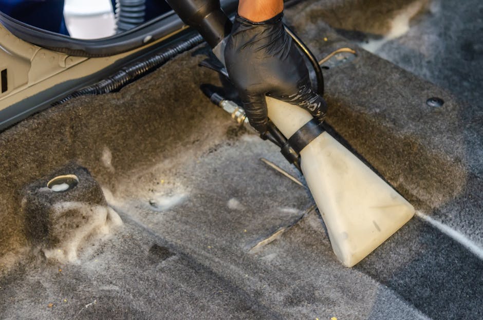 A professional cleaner dressed in a white protective suit, face mask, and blue gloves is performing deep cleaning or sanitisation work on a beige carpet in a well-lit, modern living room. The cleaner